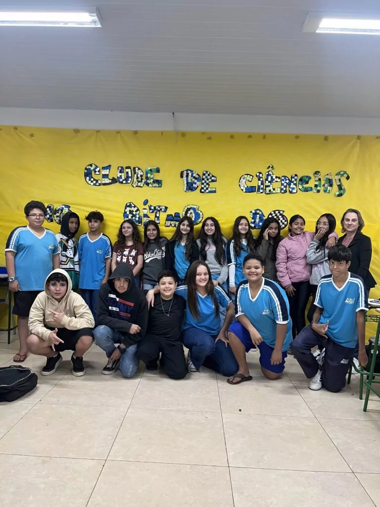 A foto é do clube de ciências ‘No Ritmo do Conhecimento’ do Colégio Estadual Doutor Camargo, na cidade de mesmo nome (Doutor Camargo). A imagem mostra um grupo de estudantes e uma professora em uma sala de aula em frente ao mural do clube “No Ritmo do Conhecimento”. As crianças estão sorrindo e posicionadas ao redor de uma mesa com cartazes coloridos e materiais escolares espalhados, indicando que participaram de uma atividade criativa ou expositiva. Ao fundo, observa-se um mural decorado com desenhos, recortes e cartazes relacionados ao projeto. A professora está de pé, junto aos alunos, e todos aparentam estar orgulhosos e felizes com o trabalho realizado. A cena transmite um ambiente de cooperação, aprendizado lúdico e valorização do conhecimento por meio de atividades em grupo.