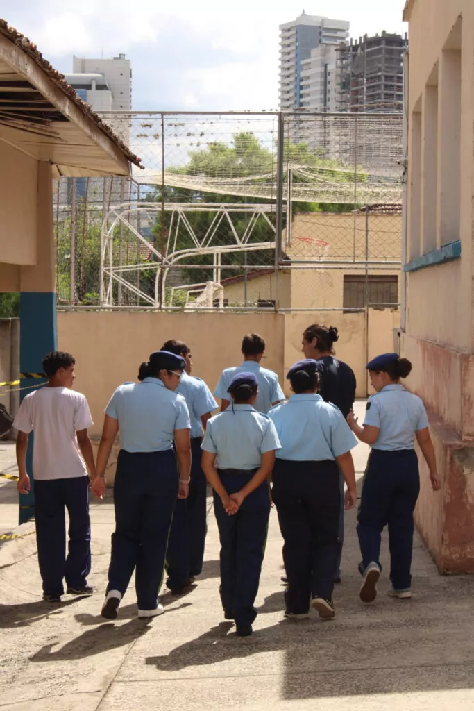 A foto mostra alguns estudantes andando enquanto conversam com um bolsista da Rede de Clubes. A sua frente é possível ver a quadra da escola com uma tabela e cesta de basquete.