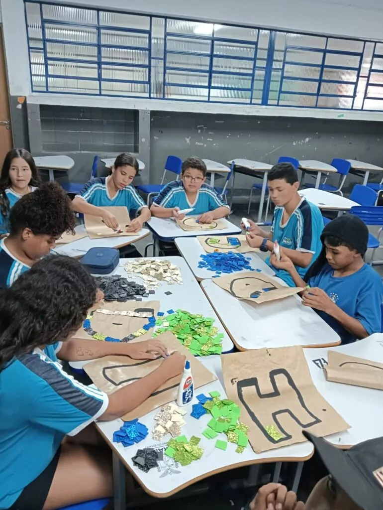 A imagem mostra uma sala de aula ampla e bem iluminada, com várias crianças sentadas em grupos ao redor de mesas retangulares. As crianças estão envolvidas em uma atividade, algumas escrevendo ou desenhando em folhas de papéis. Há materiais escolares espalhados sobre as mesas, como lápis, estojos e cadernos. Ao fundo, é possível ver murais decorativos coloridos e armários azuis com etiquetas. Também há cartazes educativos fixados nas paredes, contribuindo para um ambiente de aprendizagem estimulante. A atmosfera da sala parece alegre e organizada, sugerindo uma aula participativa e dinâmica.