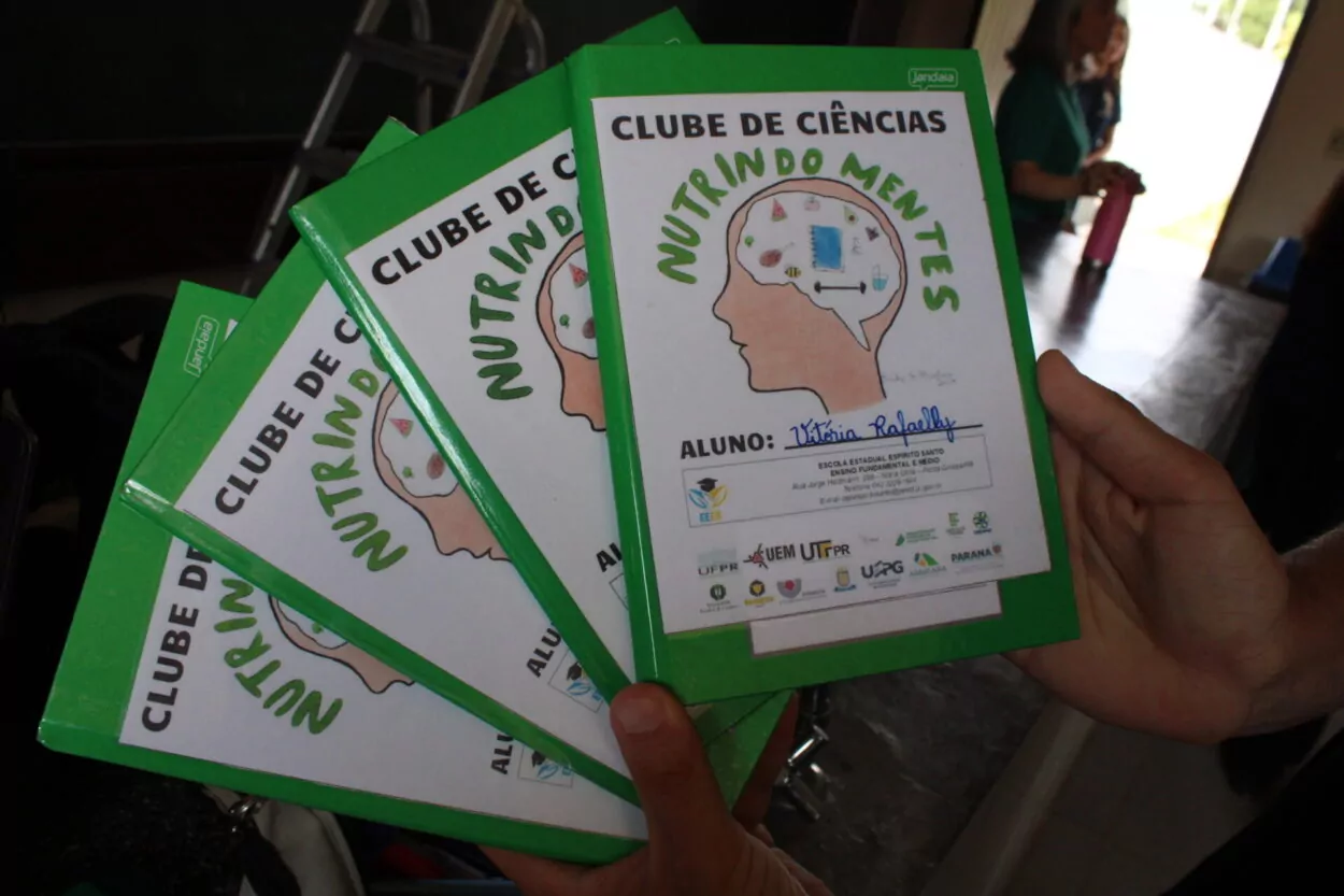 A foto mostra cadernos de capa verde com o título Clube de Ciências Nutrindo Mentes, o nome do aluno e um desenho de uma cabeça.
