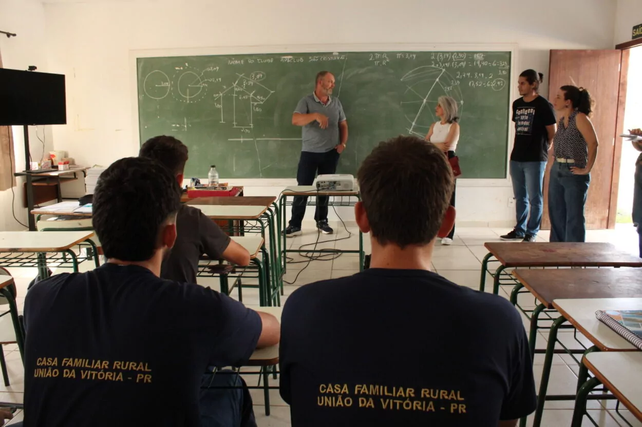 A foto mostra alunos dentro de sala de aula observando o professor conversar com a articuladora dos clubes da UEPG. Os estudantes estão de costas, de uniforme azul, no qual é possível ler o nome da Instituição em que estudam, Casa Familiar Rural de União da Vitória