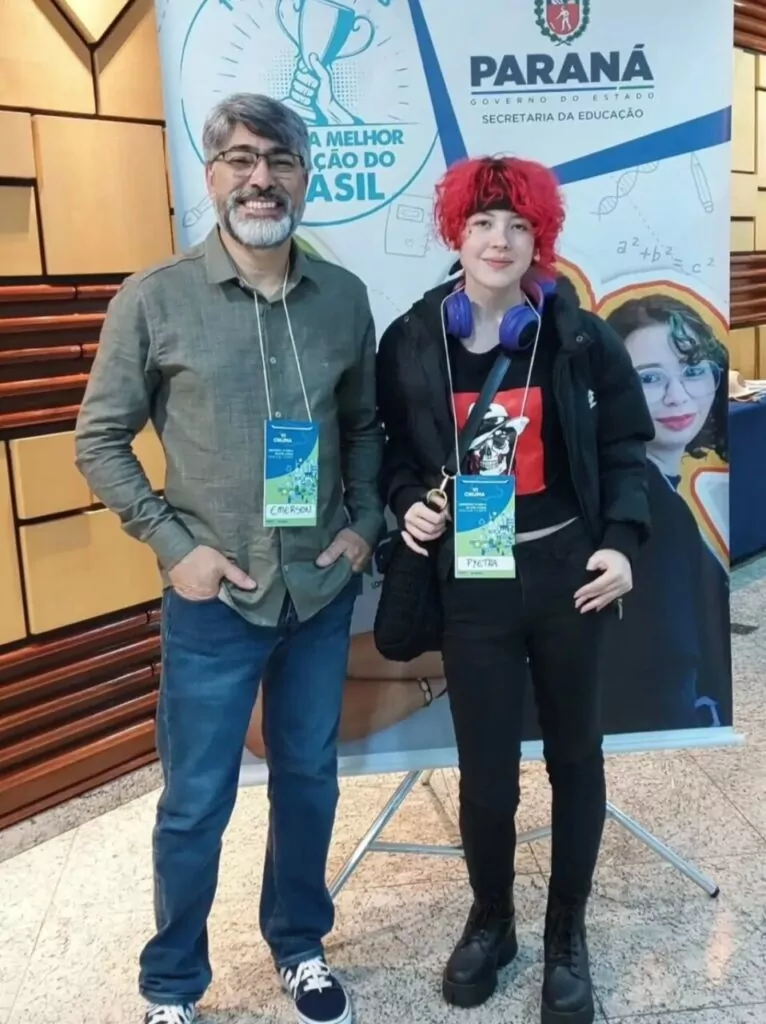 Duas pessoas estão em pé e sorrindo para a foto. À esquerda, um homem de cabelos e barba grisalhos, usa óculos, camisa cinza de manga comprida e calça jeans azul. À direita, uma jovem com cabelo curto e vermelho, veste jaqueta preta, calça preta e botas pretas. Ela tem um fone de ouvido roxo no pescoço e uma camiseta preta com estampa vermelha. Ambos usam crachás de identificação pendurados no pescoço. Ao fundo, há um banner da Secretaria da Educação do Paraná com frases e ilustrações coloridas.