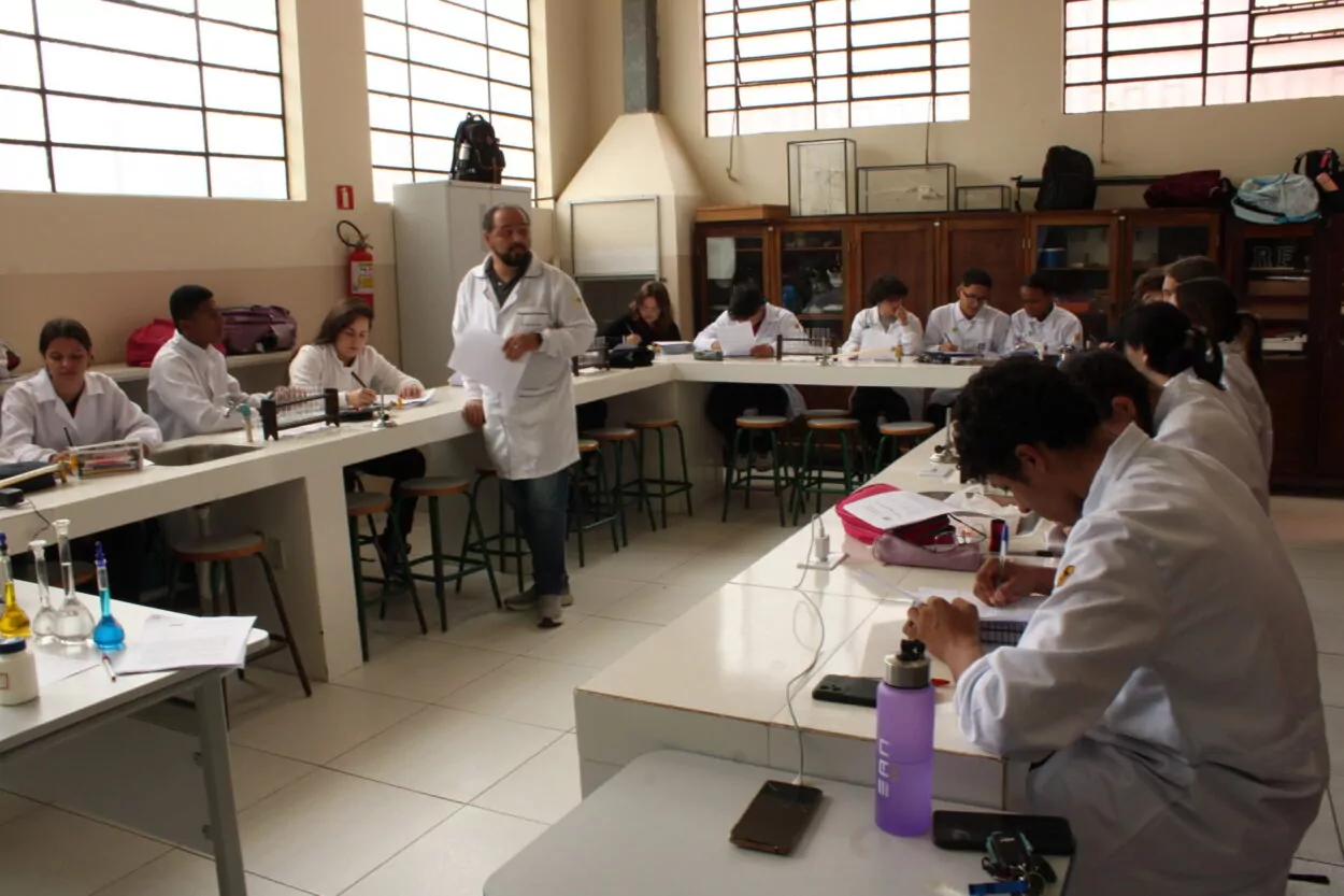 A foto mostra clubistas do Exploradores da Ciência, que participarão do 5º Paraná Faz Ciência. Eles estão em um laboratório de ciências com vidrarias e equipamentos, fazendo uma atividade em uma bancada enquanto o professor faz uma explicação. (Foto/Talula)