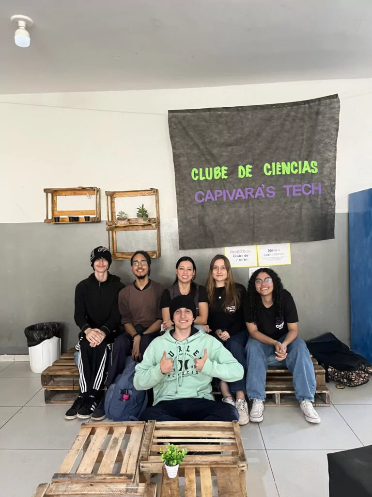 A imagem é uma galeria composta por seis fotografias, que retratam atividades desenvolvidas por diferentes grupos escolares ligados a Clubes de Ciências. A primeira foto tem um grupo de alunos sorrindo. O grupo está sentado em um ambiente interno decorado com plantas em prateleiras e cartazes. A segunda imagem mostra uma sala de informática com vários estudantes sentados em frente a computadores de mesa. Eles estão organizados em duas fileiras, cada uma voltada para o centro da sala, com monitores, teclados e fones de ouvido disponíveis. A maioria dos alunos parecem estar concentrados nas telas, digitando ou usando os fones. A sala tem iluminação fluorescente, paredes claras e alguns avisos afixados. Ao fundo, há ventiladores de parede e janelas com blocos de vidro para ventilação. O ambiente transmite uma atmosfera de estudo. A terceira fotografia tem alunos e visitantes interagindo em uma bancada comprida, realizando atividades práticas e experimentos. Há papéis com explicações e materiais sobre a mesa, indicando uma exposição interativa. A quarta foto mostra várias pessoas observando atentamente uma exposição de trabalhos em um espaço decorado com tecido verde. Na quinta foto tem um grupo de alunos apresentando um projeto do Clube de Ciências em uma sala decorada com folhas verdes penduradas. Na frente da mesa, há cartazes escritos e materiais como garrafas PET e experimentos. Na última foto há um grupo de estudantes, alguns vestidos com roupas típicas de festa junina, participando de uma atividade que envolve códigos QR. Uma das alunas segura um cartaz com o título “Quiz da ciência”, sugerindo uma abordagem lúdica para aprender conteúdos científicos.