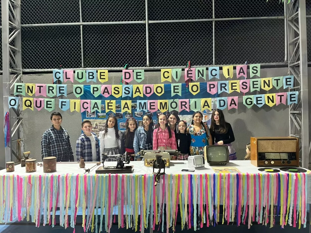 A galeria é composta por cinco fotografias, retratando diferentes momentos de atividades escolares desenvolvidas pelos Clubes de Ciências. A primeira foto expõe um grupo de estudantes e um professor posicionados atrás de três composteiras verdes com etiquetas, em um projeto que aborda o reaproveitamento de resíduos orgânicos. A segunda foto mostra alunos e professores posando ao redor de uma bancada com frascos, reagentes e uma grande bacia. A cena remete a um experimento de ciências em andamento, demonstrando práticas laboratoriais e espírito de equipe. Na terceira fotografia há um grupo de estudantes apresentando o projeto do Clube de Ciências “Entre o passado e o presente, o que fica é a memória da gente”. A exposição conta com objetos antigos e decoração com fitas coloridas, promovendo uma reflexão sobre a memória. A quarta foto mostra os alunos participando de uma atividade interativa sobre reciclagem, utilizando caixas organizadas por tipo de resíduo (plástico, papel, etc.). Crianças observam e manuseiam objetos, aprendendo sobre o descarte correto de materiais. Na última foto há dois alunos que apresentam um painel, ao lado de caixas de coleta seletiva. Uma aluna observa o espaço, que contém cartazes e materiais ligados à preservação ambiental.