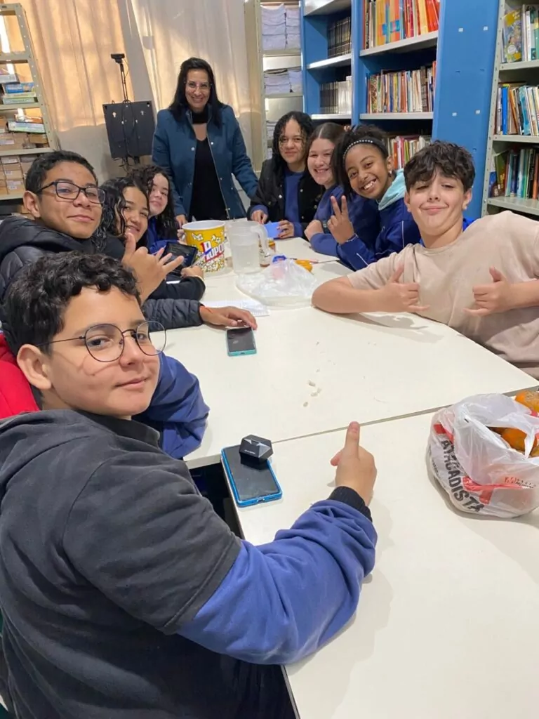 A imagem mostra um grupo de nove estudantes reunidos ao redor de uma mesa branca em uma biblioteca escolar, acompanhados por uma professora em pé ao fundo. Todos parecem felizes, sorrindo e fazendo gestos descontraídos com as mãos. A mesa está repleta de itens como um balde de pipoca, jarra com água, celular, pote de mel, sacola com laranjas e cadernos. Ao fundo, há prateleiras cheias de livros, reforçando o ambiente educativo. A cena transmite um clima de amizade, aprendizado e acolhimento.