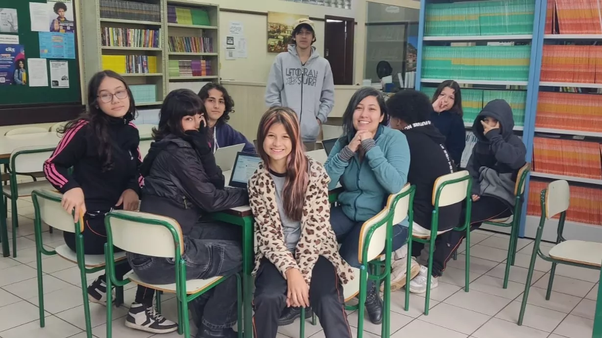 A foto mostra um grupo de nove jovens reunidos em uma sala com estantes cheias de livros coloridos ao fundo. Eles estão sentados em torno de mesas com cadeiras escolares brancas e verdes. Alguns usam notebooks, enquanto outros olham para a câmera. A atmosfera é descontraída, com expressões variadas de alegria e descontração.