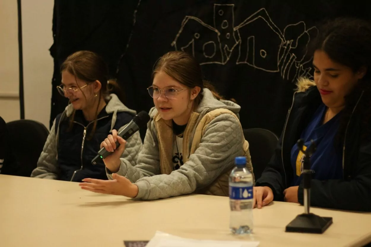 A foto mostra três jovens sentadas lado a lado atrás de uma mesa, participando de uma atividade em grupo. A jovem ao centro segura um microfone e fala com expressão atenta, enquanto faz um gesto com a mão. À esquerda, outra jovem sorri, e à direita, uma terceira olha para baixo, concentrada. Sobre a mesa há uma garrafa de água e um microfone fixo. Ao fundo, um painel escuro com desenhos de casas em linha branca decora o espaço.