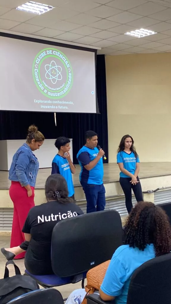 A foto mostra quatro pessoas em pé, em frente a um palco, apresentando-se para uma plateia. Três delas usam camiseta azul do Colégio Estadual João Paulo I; uma segura um microfone e fala, enquanto os outros dois escutam atentos. Ao lado, uma mulher de vestido rosa e jaqueta jeans também participa. No fundo, há um telão com o logotipo do Clube de Ciências Conexão e Sustentabilidade e a frase “Explorando conhecimento, inovando o futuro”. Algumas pessoas estão sentadas na plateia, de costas para a câmera.