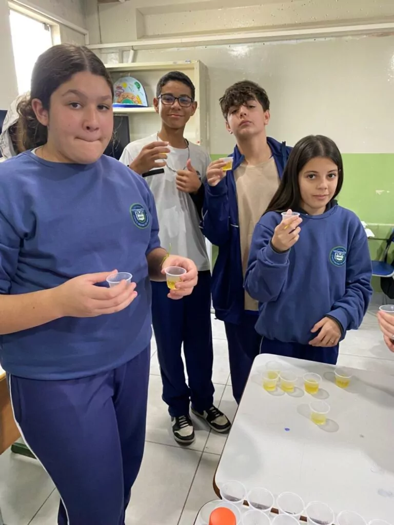 A galeria de fotos mostra momentos da apresentação e degustação do energético produzido pelo clube de ciências Ciência na Mesa. As imagens mostram estudantes na sala de aula com as mesas organizadas para oferecer o energético em copo descartáveis. Todos parecem felizes, sorrindo e curiosos com a proposta de bebida alternativa