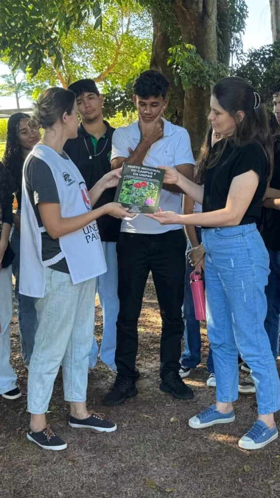 Na imagem é possível o grupo de alunos prestando atenção na explicação da professora sobre o projeto Horto Medicinal da Unipar (Universidade Paranaense de Umuarama), em viagem os alunos aproveitaram para se aprofundar no assunto e conhecer mais sobre as ervas, voltado ao estudo, cultivo e preservação de plantas com propriedades fitoterápicas. O grupo de alunos está ao ar livre e a professora responsável mostrando as amostras das plantas para que os alunos conheçam suas diferenças. É possível notar o entusiasmo entre os alunos clubistas que puderam aproximar teoria e prática.