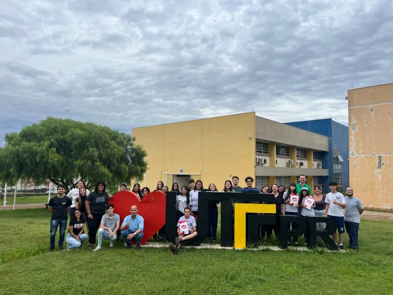 A foto mostra um grande grupo de pessoas reunidas em frente ao letreiro da UTFPR no campus de Apucarana. O letreiro é formado por um coração vermelho seguido das letras em preto e amarelo "UTFPR". O grupo é diverso, com jovens e adultos, alguns usando camisetas da UTFPR e outros segurando pastas. Ao fundo, há prédios da universidade em tons de amarelo e azul, além de uma grande árvore à esquerda. O céu está nublado.