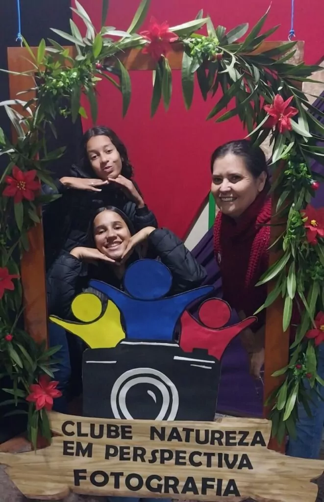A primeira fotografia é do Clube Natureza em Perspectiva. Na imagem há três pessoas — duas alunas e uma mulher adulta — que posam sorridentes em um painel decorado com folhas verdes e flores vermelhas. A moldura decorativa envolve o cartaz do Clube Natureza em Perspectiva – Fotografia, que possui um símbolo colorido com silhuetas humanas e uma câmera fotográfica estilizada. A segunda fotografia é do clube “Academia de Jovens Inventores”. Na imagem tem um grupo de estudantes reunidos em uma sala de aula. Um aluno em destaque demonstra o uso de um equipamento diante dos colegas. A mesa está repleta de materiais escolares e científicos, e o ambiente é iluminado por uma janela com cortinas claras. Ao fundo, há um cartaz indicando a presença de um clube escolar.