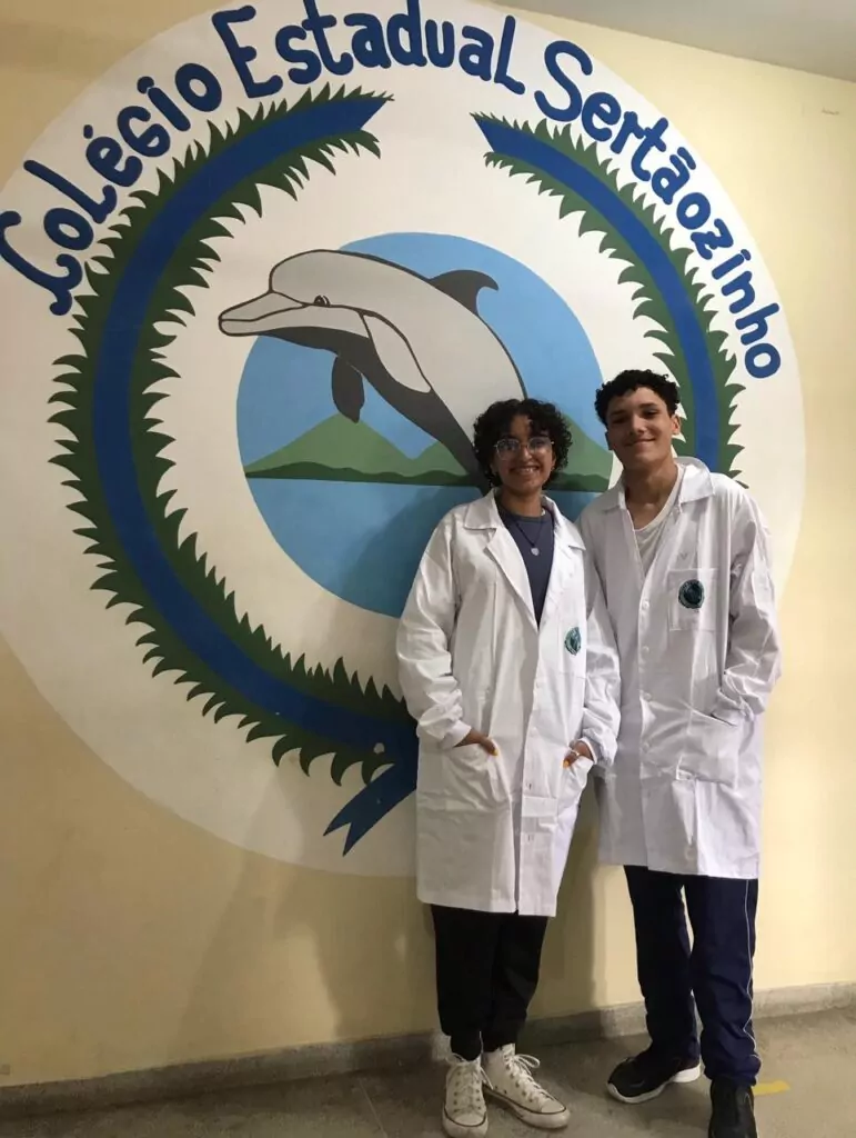 Na imagem, dois estudantes do Colégio Estadual Cívico-Militar Sertãozinho, em Matinhos (PR), posam sorridentes em frente ao mural da escola, que exibe o logotipo com um golfinho e o nome da instituição. Ambos vestem jalecos brancos com o emblema do colégio, representando o Clube de Ciências Ecocientistas.