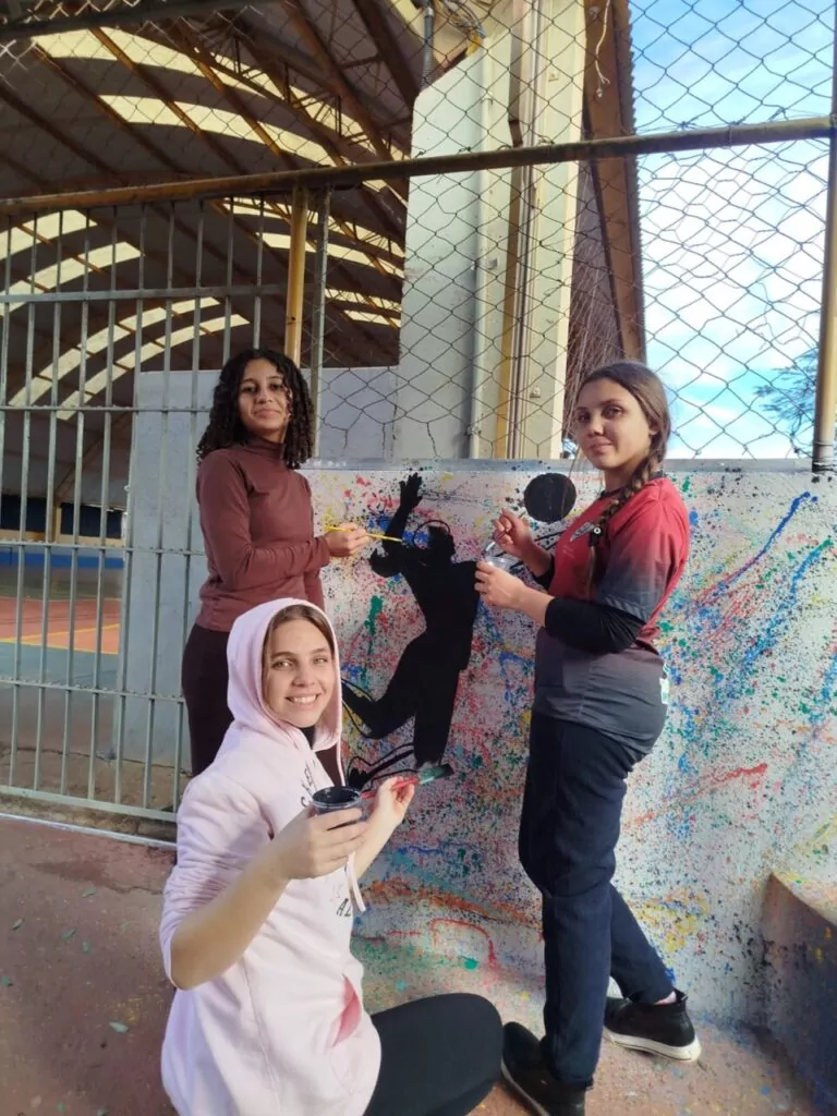 Na primeira fotografia temos a foto do clube “Tenda Cultura Viva”. A imagem mostra três jovens em uma sala de aula, posicionadas ao lado de uma televisão que exibe uma apresentação. Na tela aparece a imagem de um rosto pintado, acompanhada do texto: “Tenda Cultura Viva - Projeto Paraná Faz Ciência”. As três jovens sorriem para a foto, usando crachás pendurados no pescoço. A sala possui um quadro verde ao fundo, com um desenho de flor, e armários metálicos próximos à janela, por onde entra bastante luz natural através de uma cortina clara. A segunda fotografia é do Clube “Paisagismo Sustentável”. A imagem mostra três estudantes participando de uma atividade de pintura em um muro. O muro está coberto por respingos coloridos de tinta e, no centro, há a figura de uma pessoa jogando vôlei. As alunas estão com pincéis e potes de tinta nas mãos, sorrindo e interagindo com a arte. Ao fundo, aparece a estrutura de uma quadra esportiva coberta, cercada por grades e tela de proteção. Enquanto a terceira fotografia é do clube “Green Lab”. A imagem mostra uma sala decorada com fitas coloridas penduradas no teto. No centro, um grupo de crianças, muitas delas usando casacos e moletons, está reunido de frente para uma apresentação. Na parte da frente da sala, há quatro jovens responsáveis pela atividade: dois estão em pé e dois sentados atrás de uma mesa coberta com uma toalha azul, onde há garrafas e materiais organizados. As paredes têm cartazes com fotos e ilustrações, além de símbolos relacionados à ciência.