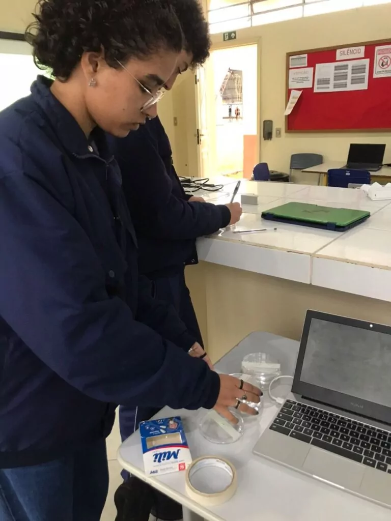 Na imagem, uma estudante do Colégio Estadual Cívico-Militar Sertãozinho, em Matinhos (PR), realiza uma atividade prática no laboratório escolar. Ela manipula placas de Petri sobre a bancada, utilizando fita adesiva e materiais de higiene, enquanto outro estudante anota observações ao lado. Um notebook aberto auxilia o registro das etapas do experimento.