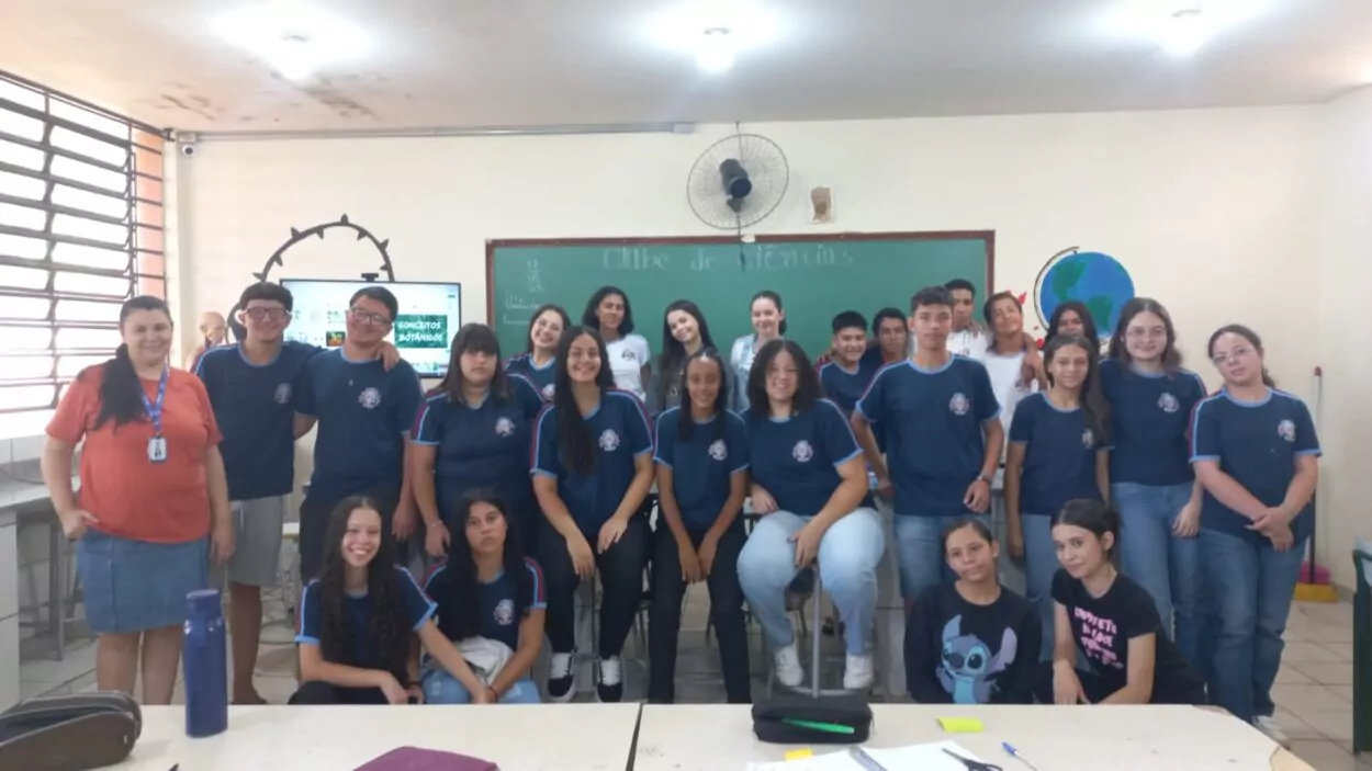 A primeira imagem mostra um grupo de estudantes e uma professora em uma sala de aula em frente ao mural do clube “No Ritmo do Conhecimento”. As crianças estão sorrindo e posicionadas ao redor de uma mesa com cartazes coloridos e materiais escolares espalhados. A segunda imagem mostra um grupo de estudantes posando para uma foto dentro de uma sala de aula. O grupo é formado por 14 alunos e uma adulta, a professora. Eles estão dispostos em duas fileiras: a primeira sentada e a segunda em pé. Atrás deles há um mural com cartazes e trabalhos escolares expostos na parede. A terceira foto mostra um grupo de estudantes e uma professora reunidos em uma sala de aula. Eles estão posando para uma foto em frente a um quadro verde, no qual está escrito “Clube de Ciências”. No ambiente, é possível ver carteiras na frente, uma televisão ligada exibindo a palavra “Conceitos Botânicos” e uma janela com grades à esquerda. A quarta fotografia é do Clube Jovens Cientistas Kennedy. A imagem mostra um grupo grande de estudantes posando para a foto em um pátio externo, com árvores de folhagem vermelha ao fundo e um muro branco atrás deles. Algumas alunas estão agachadas na frente, junto com duas mulheres adultas, enquanto o restante da turma permanece em pé. Um homem adulto está à esquerda do grupo. A quinta fotografia mostra um grupo de estudantes reunidos em uma sala de aula, participando do Clube de Ciência. Eles vestem camisetas verdes do colégio e estão organizados ao redor de uma mesa de laboratório. Ao fundo, há uma tela que apresenta o tema do projeto: “Plant Scientists Club: Descobrindo alternativas no combate de plantas daninhas a partir do estudo de bioinsumos”. Na sexta imagem, um grupo de estudantes e professores está reunido em um espaço externo, em frente a árvores e cercas metálicas. Eles posam sorridentes para a foto. Na frente, duas mulheres seguram uma bandeira verde e branca com o desenho de uma capivara e os dizeres “CAPCIÊNCIA – Clube de Ciências”.