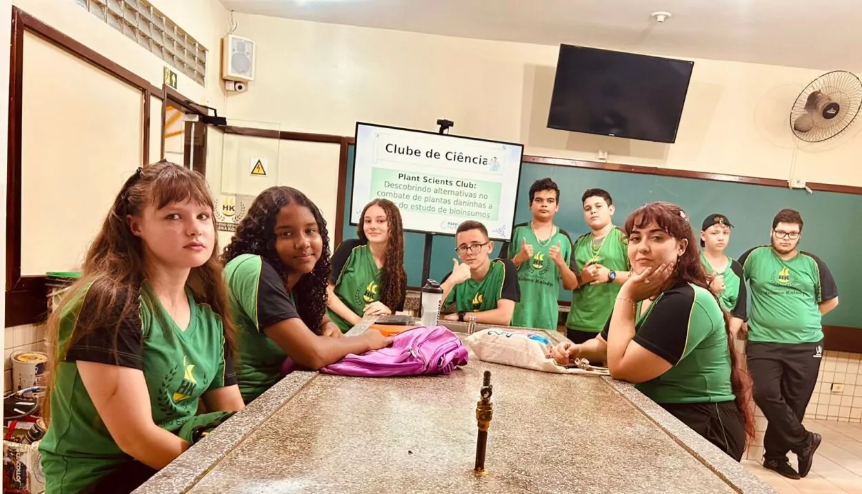 A primeira imagem mostra um grupo de estudantes e uma professora em uma sala de aula em frente ao mural do clube “No Ritmo do Conhecimento”. As crianças estão sorrindo e posicionadas ao redor de uma mesa com cartazes coloridos e materiais escolares espalhados. A segunda imagem mostra um grupo de estudantes posando para uma foto dentro de uma sala de aula. O grupo é formado por 14 alunos e uma adulta, a professora. Eles estão dispostos em duas fileiras: a primeira sentada e a segunda em pé. Atrás deles há um mural com cartazes e trabalhos escolares expostos na parede. A terceira foto mostra um grupo de estudantes e uma professora reunidos em uma sala de aula. Eles estão posando para uma foto em frente a um quadro verde, no qual está escrito “Clube de Ciências”. No ambiente, é possível ver carteiras na frente, uma televisão ligada exibindo a palavra “Conceitos Botânicos” e uma janela com grades à esquerda. A quarta fotografia é do Clube Jovens Cientistas Kennedy. A imagem mostra um grupo grande de estudantes posando para a foto em um pátio externo, com árvores de folhagem vermelha ao fundo e um muro branco atrás deles. Algumas alunas estão agachadas na frente, junto com duas mulheres adultas, enquanto o restante da turma permanece em pé. Um homem adulto está à esquerda do grupo. A quinta fotografia mostra um grupo de estudantes reunidos em uma sala de aula, participando do Clube de Ciência. Eles vestem camisetas verdes do colégio e estão organizados ao redor de uma mesa de laboratório. Ao fundo, há uma tela que apresenta o tema do projeto: “Plant Scientists Club: Descobrindo alternativas no combate de plantas daninhas a partir do estudo de bioinsumos”. Na sexta imagem, um grupo de estudantes e professores está reunido em um espaço externo, em frente a árvores e cercas metálicas. Eles posam sorridentes para a foto. Na frente, duas mulheres seguram uma bandeira verde e branca com o desenho de uma capivara e os dizeres “CAPCIÊNCIA – Clube de Ciências”.