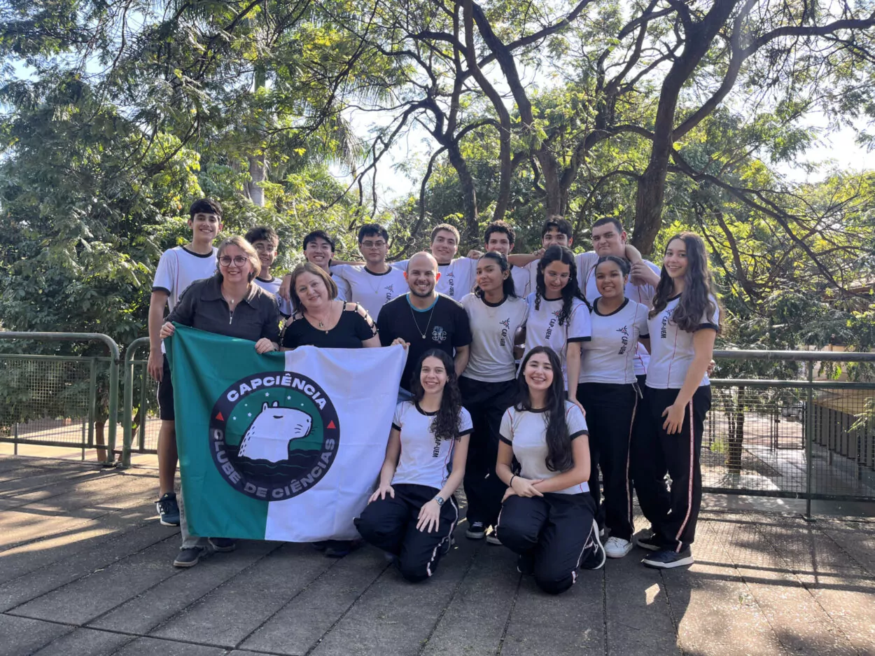 A primeira imagem mostra um grupo de estudantes e uma professora em uma sala de aula em frente ao mural do clube “No Ritmo do Conhecimento”. As crianças estão sorrindo e posicionadas ao redor de uma mesa com cartazes coloridos e materiais escolares espalhados. A segunda imagem mostra um grupo de estudantes posando para uma foto dentro de uma sala de aula. O grupo é formado por 14 alunos e uma adulta, a professora. Eles estão dispostos em duas fileiras: a primeira sentada e a segunda em pé. Atrás deles há um mural com cartazes e trabalhos escolares expostos na parede. A terceira foto mostra um grupo de estudantes e uma professora reunidos em uma sala de aula. Eles estão posando para uma foto em frente a um quadro verde, no qual está escrito “Clube de Ciências”. No ambiente, é possível ver carteiras na frente, uma televisão ligada exibindo a palavra “Conceitos Botânicos” e uma janela com grades à esquerda. A quarta fotografia é do Clube Jovens Cientistas Kennedy. A imagem mostra um grupo grande de estudantes posando para a foto em um pátio externo, com árvores de folhagem vermelha ao fundo e um muro branco atrás deles. Algumas alunas estão agachadas na frente, junto com duas mulheres adultas, enquanto o restante da turma permanece em pé. Um homem adulto está à esquerda do grupo. A quinta fotografia mostra um grupo de estudantes reunidos em uma sala de aula, participando do Clube de Ciência. Eles vestem camisetas verdes do colégio e estão organizados ao redor de uma mesa de laboratório. Ao fundo, há uma tela que apresenta o tema do projeto: “Plant Scientists Club: Descobrindo alternativas no combate de plantas daninhas a partir do estudo de bioinsumos”. Na sexta imagem, um grupo de estudantes e professores está reunido em um espaço externo, em frente a árvores e cercas metálicas. Eles posam sorridentes para a foto. Na frente, duas mulheres seguram uma bandeira verde e branca com o desenho de uma capivara e os dizeres “CAPCIÊNCIA – Clube de Ciências”.