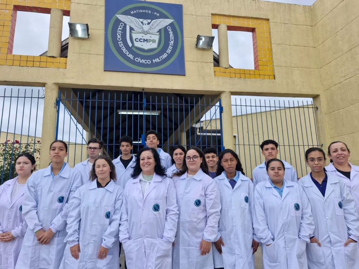 Grupo de estudantes e professoras do Colégio Estadual Cívico-Militar Sertãozinho, em Matinhos (PR), posa em frente ao portão da escola. Todos vestem jalecos brancos com o emblema do colégio, simbolizando a participação no Clube de Ciências Ecocientistas. Ao fundo, é possível ver o prédio da instituição e o brasão do colégio acima do portão azul.