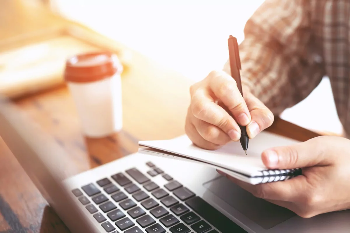 A imagem mostra uma cena de estudo. Em primeiro plano, vemos uma mão segurando uma caneta preta e escrevendo em um caderno de folhas brancas com espiral lateral. O caderno está apoiado parcialmente sobre um notebook prateado, que está aberto.