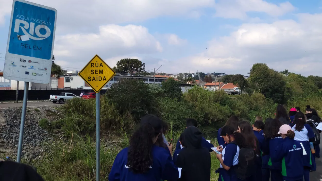 Um grupo de estudantes caminha em fila às margens do rio Belém, vestidos com uniformes azul-marinho com detalhes em verde e a inscrição “Paraná Integral” nas costas. Eles seguram pranchetas e fazem anotações enquanto observam o ambiente ao redor. À esquerda da imagem, em primeiro plano, aparecem duas placas de trânsito: uma azul e branca com os dizeres “Aqui passa o rio Belém” e outra amarela em formato losangular escrito “Rua sem saída”. Ao fundo, é possível ver casas de telhado vermelho, árvores e um céu parcialmente nublado. A cena transmite a ideia de investigação em campo, com os alunos registrando observações sobre o território.