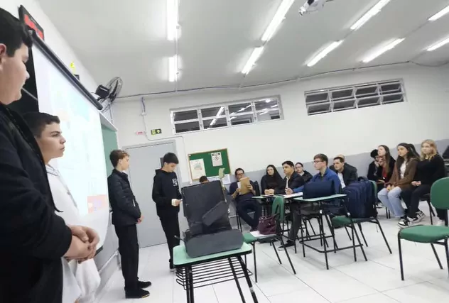 A foto mostra os quatro estudantes apresentando o trabalho premiado, eles estão em uma sala de aula, ao lado de uma projeção em tela. A sua frente, sentados em cadeiras verdes, estudantes assistem atentos a comunicação. Ao centro também o Robô Papa-Pilhas é exibido em uma carteira.
