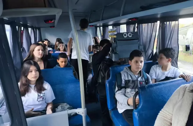 A foto mostra estudantes dentro de um ônibus com assentos azuis. Eles olham para a frente, com expressões neutras. Um dos estudantes está de pé no corredor, andando para o fundo. Um clubista também segura o Robô Papa-Pilhas