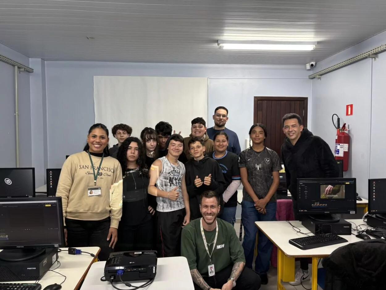 Um grupo de treze pessoas está reunido em uma sala de computadores. Eles estão posicionados em pé e sorrindo para a foto, enquanto um homem, agachado à frente, também sorri. À esquerda, uma mulher com moletom bege e crachá aparece de pé, e ao fundo há jovens e adultos lado a lado. À direita, outro homem veste um casaco preto. Nas mesas ao redor, é possível ver monitores de computador e um projetor. A parede da sala é clara, com um extintor vermelho fixado próximo à porta de madeira.