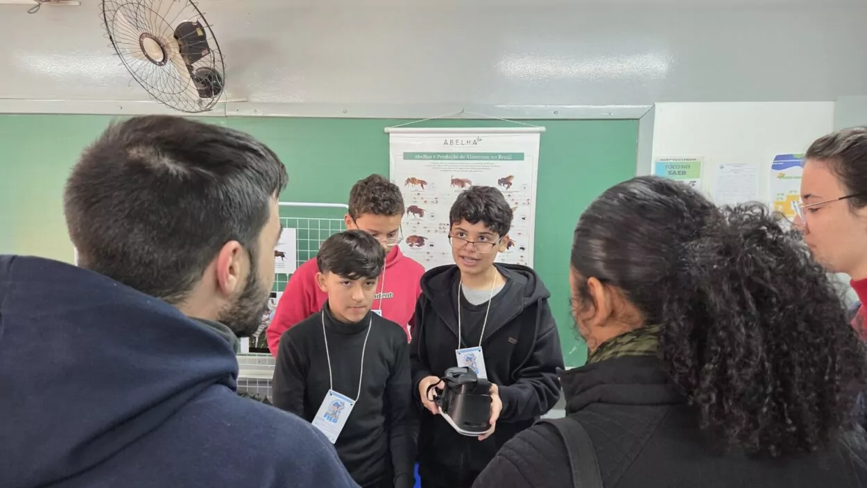 Na imagem, alguns estudantes apresentam um projeto em uma sala de aula. Dois deles estão à frente, um segurando um equipamento de realidade virtual, enquanto outras pessoas observam atentamente. Ao fundo, há cartazes educativos fixados na parede verde.