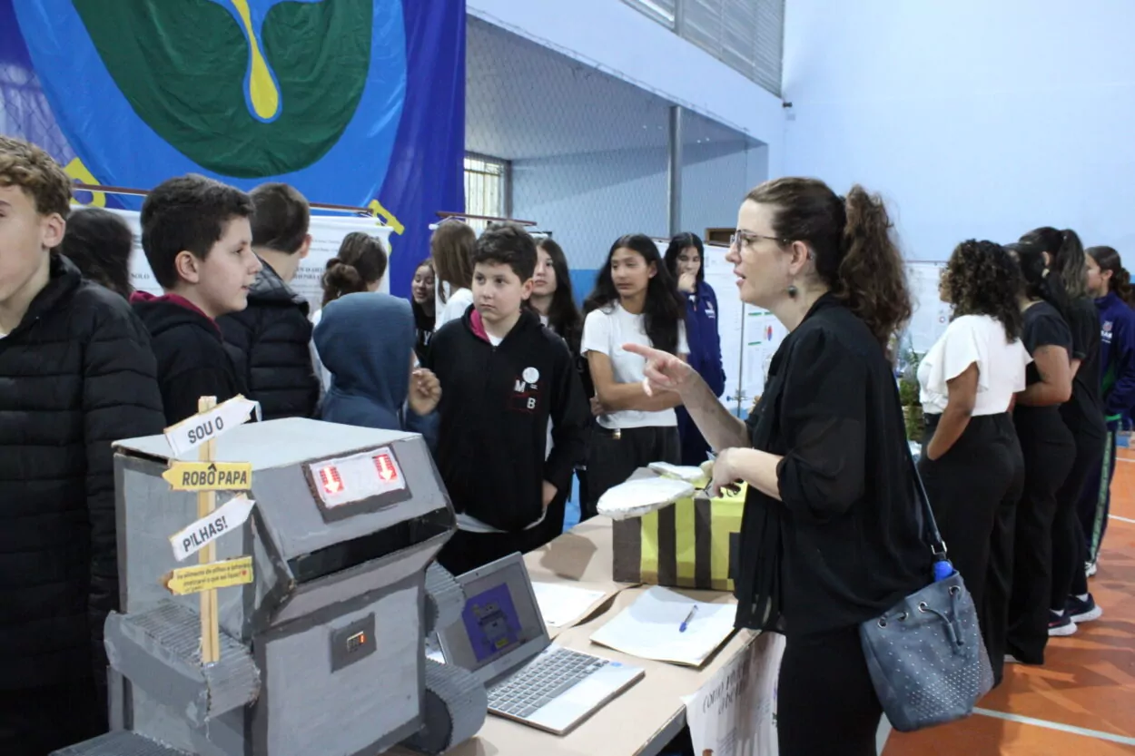 A foto mostra vários alunos em uma Feira de Ciências, entre estudantes observando e apresentando seus trabalhos. O ambiente é fechado, há uma bandeira com o símbolo das ciências biológicas ao fundo, e banners enfileirados também ao fundo. Em primeiro plano, uma professora conversa com os clubistas do Pilha nas Abelhas, entre eles, uma mesa com materiais expostos, incluindo o Robô Papa Pilhas. Feito de caixas pintadas e placas de led em forma de seta que apontam para sua boca, o robô é um projeto do clube para incentivo do descarte adequado de pilhas e baterias.