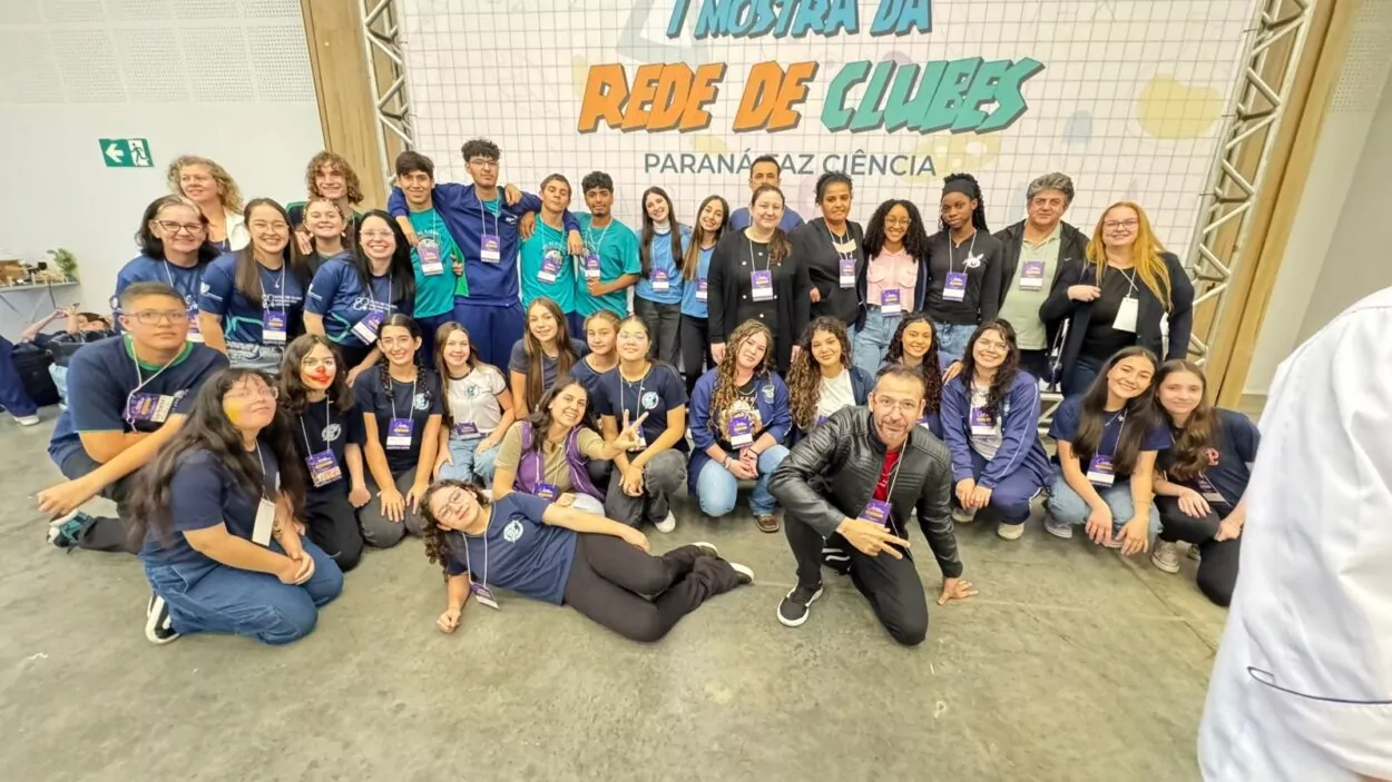 Na foto apresentada, é possível ver no fundo da imagem atrás dos alunos e professores, em destaque, a I Mostra da Rede de Clubes Paraná FAZ Ciência, todos celebram a participação no dia da feira, depois de um dia muito produtivo.