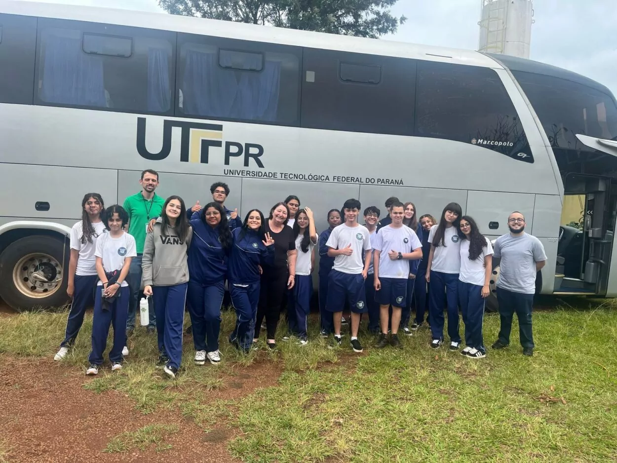 A foto mostra um grupo de estudantes e professores posando em frente a um ônibus branco e cinza da Universidade Tecnológica Federal do Paraná (UTFPR). O ônibus está estacionado sobre um gramado, e o logotipo da universidade aparece em destaque na lateral. As pessoas estão sorrindo e fazem gestos de alegria, como joinhas e acenos. A maioria veste uniformes escolares azuis e brancos; duas pessoas usam roupas casuais. O clima parece nublado, e há vegetação e uma estrutura alta ao fundo.