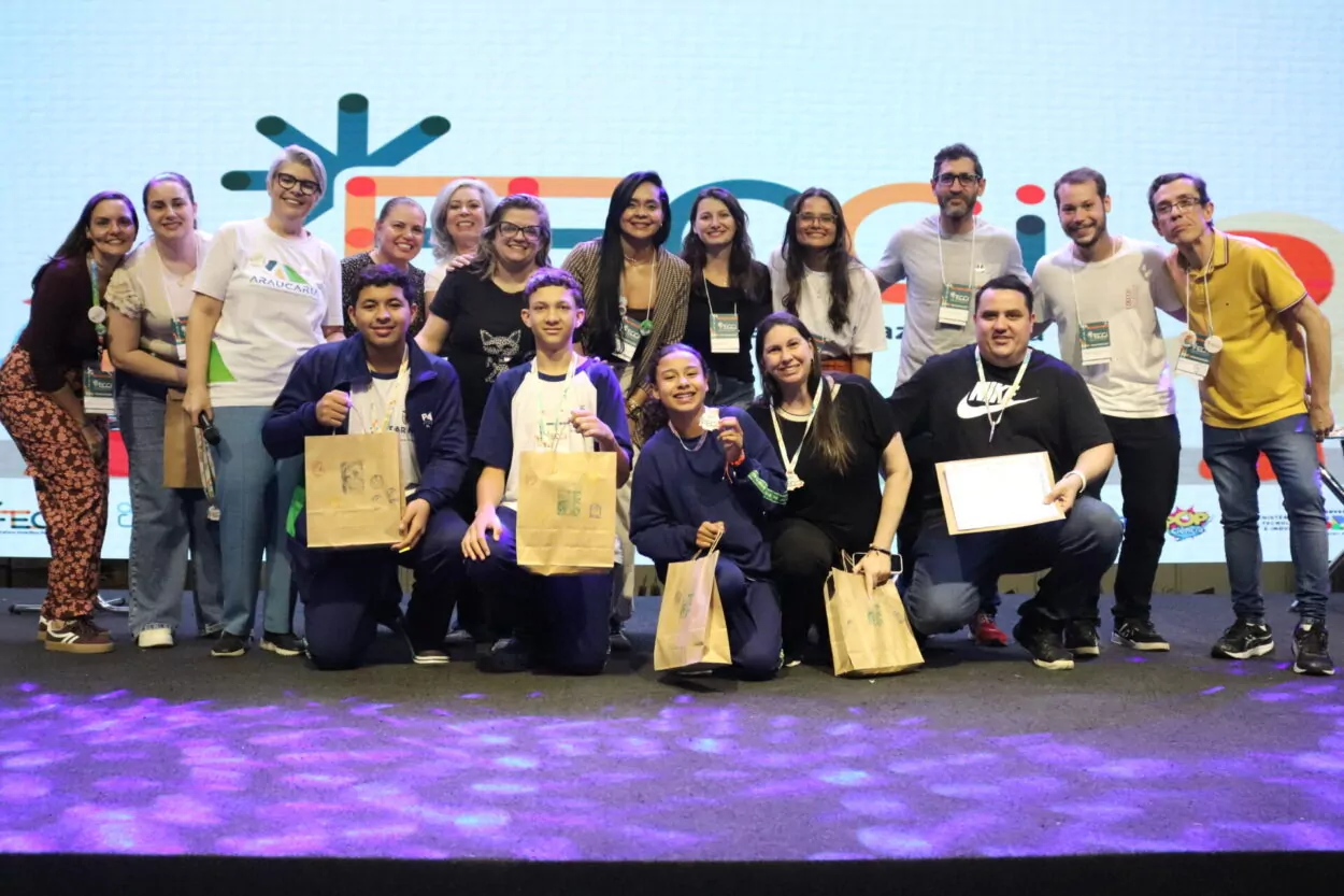 A primeira imagem é do Clube Show da Química do Colégio Estadual Cristóvão Colombo, de Jardim Alegre. A imagem mostra um grupo de pessoas posando sorridentes em um palco. Há cerca de dezesseis pessoas organizadas em duas fileiras, com alguns agachados na frente e outros em pé atrás. Os jovens na frente seguram sacolas de papel e certificados. Na segunda imagem vemos o Clube Entre o Passado e o Presente o Que Fica é a Memória da Gente do Colégio Estadual Campo D. Pedro I de Lidianópolis. A imagem mostra um grupo de pessoas reunidas em um palco durante um evento. Ao todo, há cerca de quinze pessoas, entre jovens e adultos, divididas em duas fileiras. As pessoas na frente seguram certificados e sacolas de papel. No fundo, há um grande telão colorido com o logotipo do evento.
