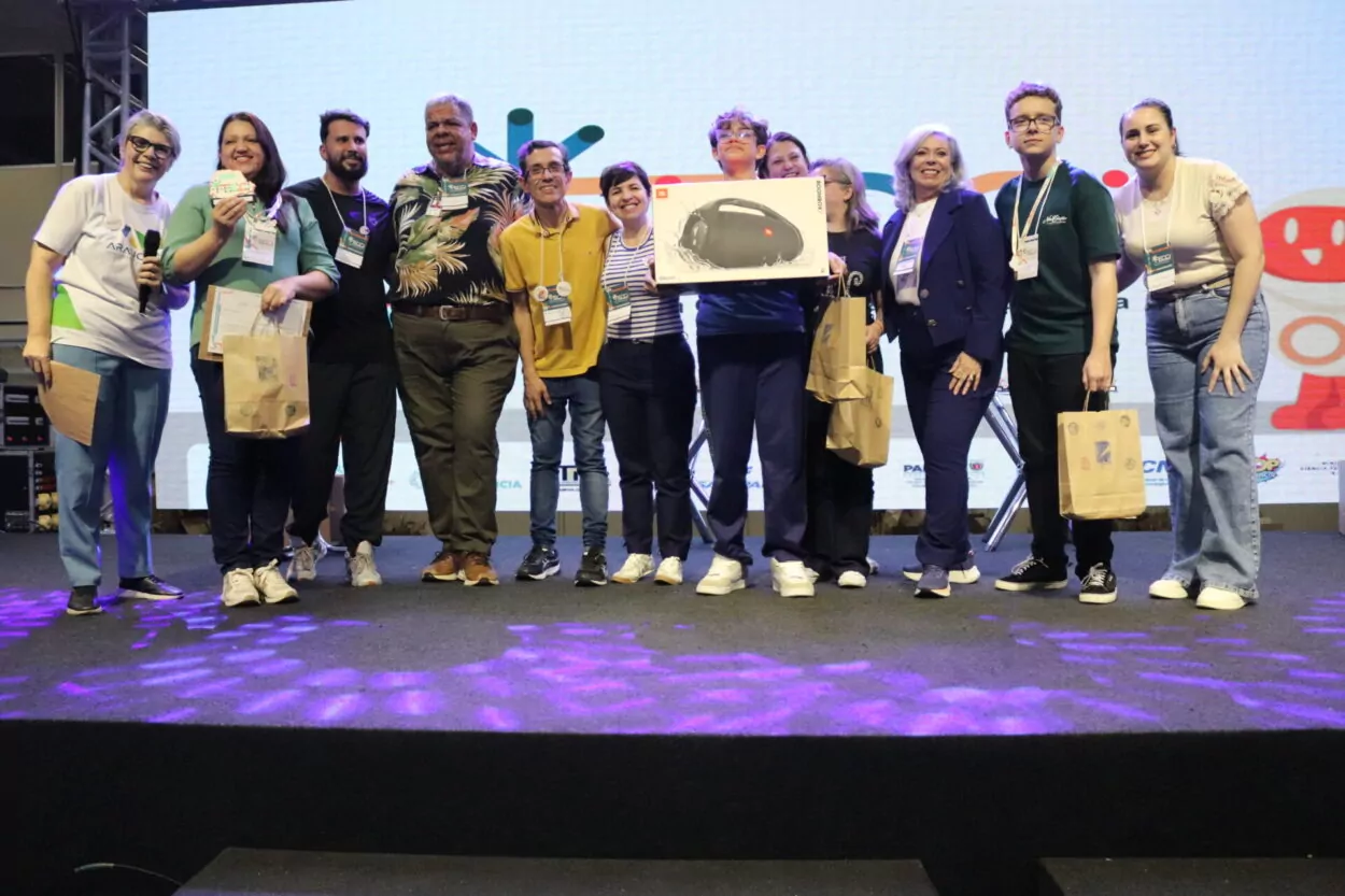 A primeira imagem é do Clube de Ciências Tenda Cultura Viva, do Colégio Estadual Professora Hilda Trautwein Kamal. Na foto há um grupo de pessoas reunidas sobre um palco, sorrindo e posando para a foto durante um evento. Ao fundo, há um telão sugerindo um encontro educacional. Os participantes estão lado a lado, segurando certificados, sacolas de papel e prêmios. Uma pessoa ao centro segura uma grande caixa de uma caixa de som JBL. Todos usam crachás de identificação e vestem roupas casuais. Na segunda fotografia vemos o Dengue Bot, do Colégio Estadual Vereador José Balan. Um grupo grande de pessoas reunidas sobre um palco, sorrindo para uma foto coletiva. Ao fundo, há um telão iluminado com elementos gráficos coloridos, sugerindo que o evento é de caráter educacional. Na parte da frente, alguns jovens seguram sacolas de papel e usam crachás de identificação e camisetas com o logotipo do Paraná. O Paisagismo Sustentável da Escola Estadual Manuel Bandeira, de Alto Piquiri, é a terceira foto. A imagem mostra um grupo numeroso de pessoas reunidas sobre um palco, posando para uma foto durante um evento educacional. Ao fundo, há um telão iluminado com elementos gráficos, reforçando o clima festivo e institucional. Na parte da frente, alguns jovens e adultos exibem certificados, troféus e prêmios. A quarta foto é do Clube Green Lab do Colégio Estadual Almirante Tamandaré, de Cruzeiro do Oeste. Na imagem é possível ver pessoas posando juntas em um palco. Há cerca de quinze pessoas, entre adultos e jovens, algumas agachadas na frente e outras em pé atrás. Os jovens na frente seguram certificados e sacolas de papel. No fundo, há um telão grande com um logotipo colorido.
