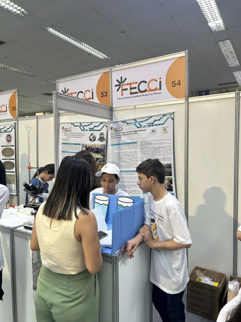 A imagem mostra um estande de feira científica identificado como FECCI– Feira de Cultura Científica Paraná Faz Ciência, com os números 53 e 54 no topo das estruturas. Dois estudantes, que parecem estar apresentando um projeto. Eles conversam com uma visitante que está de costas, vestindo calça verde e blusa clara. Sobre a mesa há uma maquete ou modelo didático com desenhos de recipientes de água, sugerindo que o projeto aborda algum tema relacionado a recursos hídricos, qualidade da água ou processos científicos. Ao fundo, há um pôster grande com textos, imagens e gráficos explicando o experimento.