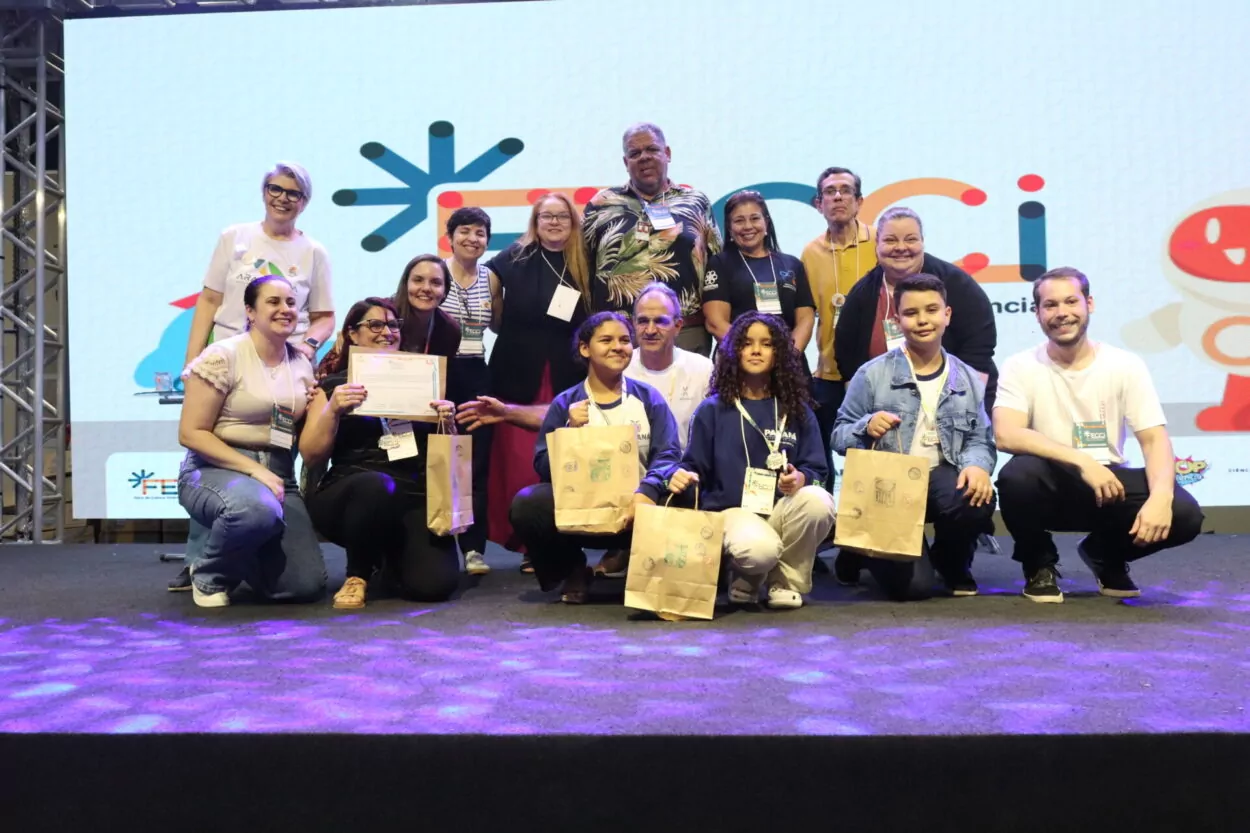 A primeira imagem é do Clube Show da Química do Colégio Estadual Cristóvão Colombo, de Jardim Alegre. A imagem mostra um grupo de pessoas posando sorridentes em um palco. Há cerca de dezesseis pessoas organizadas em duas fileiras, com alguns agachados na frente e outros em pé atrás. Os jovens na frente seguram sacolas de papel e certificados. Na segunda imagem vemos o Clube Entre o Passado e o Presente o Que Fica é a Memória da Gente do Colégio Estadual Campo D. Pedro I de Lidianópolis. A imagem mostra um grupo de pessoas reunidas em um palco durante um evento. Ao todo, há cerca de quinze pessoas, entre jovens e adultos, divididas em duas fileiras. As pessoas na frente seguram certificados e sacolas de papel. No fundo, há um grande telão colorido com o logotipo do evento.