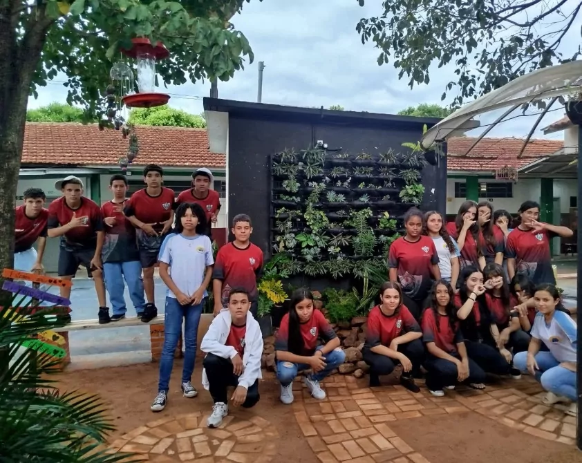 Na primeira fotografia é possível ver um grupo de pessoas posando juntas dentro de uma sala de aula. Ao todo, são dez pessoas: oito jovens e dois adultos. Eles estão posicionados lado a lado. O ambiente é típico de sala de aula, com paredes amarelas, quadro verde ao fundo e piso de cerâmica. A segunda imagem é do Clube Ilha da Ciência. A imagem mostra um grupo de estudantes e duas mulheres posando em um ambiente ao ar livre, com árvores, arbustos e canteiros de horta ao fundo. O grupo está sorridente, alguns em pé e outros agachados na frente. Na terceira fotografia tem um grupo de estudantes reunidos em um ambiente escolar ao ar livre, em frente a um jardim vertical com diversas plantas verdes. Eles estão distribuídos em pé e agachados, posando para a foto. A quarta imagem mostra um grupo de estudantes e um adulto reunidos em um espaço ao ar livre, posando para uma foto em frente a uma grande escultura de uma figura humana com os braços abertos. O grupo está distribuído sobre um pequeno morro coberto de grama, alguns sentados e outros em pé. A quinta imagem é do Clube Experimentando a Ciência: Descobrindo cores e novos sabores. A imagem mostra um grupo de estudantes e professores posando para uma foto ao ar livre, em frente a uma placa de madeira com o desenho de uma abelha e a palavra “Meliponário”. Estão sorridentes e organizados ao redor da placa. Na sexta imagem mostra um grupo grande de pessoas, formado por adolescentes e um adulto, posando para uma foto em uma sala. Eles estão organizados em duas fileiras: alguns sentados ou agachados na frente. A sala tem paredes claras, ventiladores fixados na parede e um quadro branco ao fundo. À esquerda, há uma mesa com uma toalha vermelha e alguns objetos decorativos para um evento escolar. A última fotografia tem um grupo de estudantes e alguns adultos reunidos para uma foto em um ambiente escolar externo. Eles estão em frente a uma parede branca com detalhes verdes e uma janela comprida com grades. O grupo está posicionado em duas fileiras, com alguns agachados na frente e outros em pé atrás, todos sorrindo.
