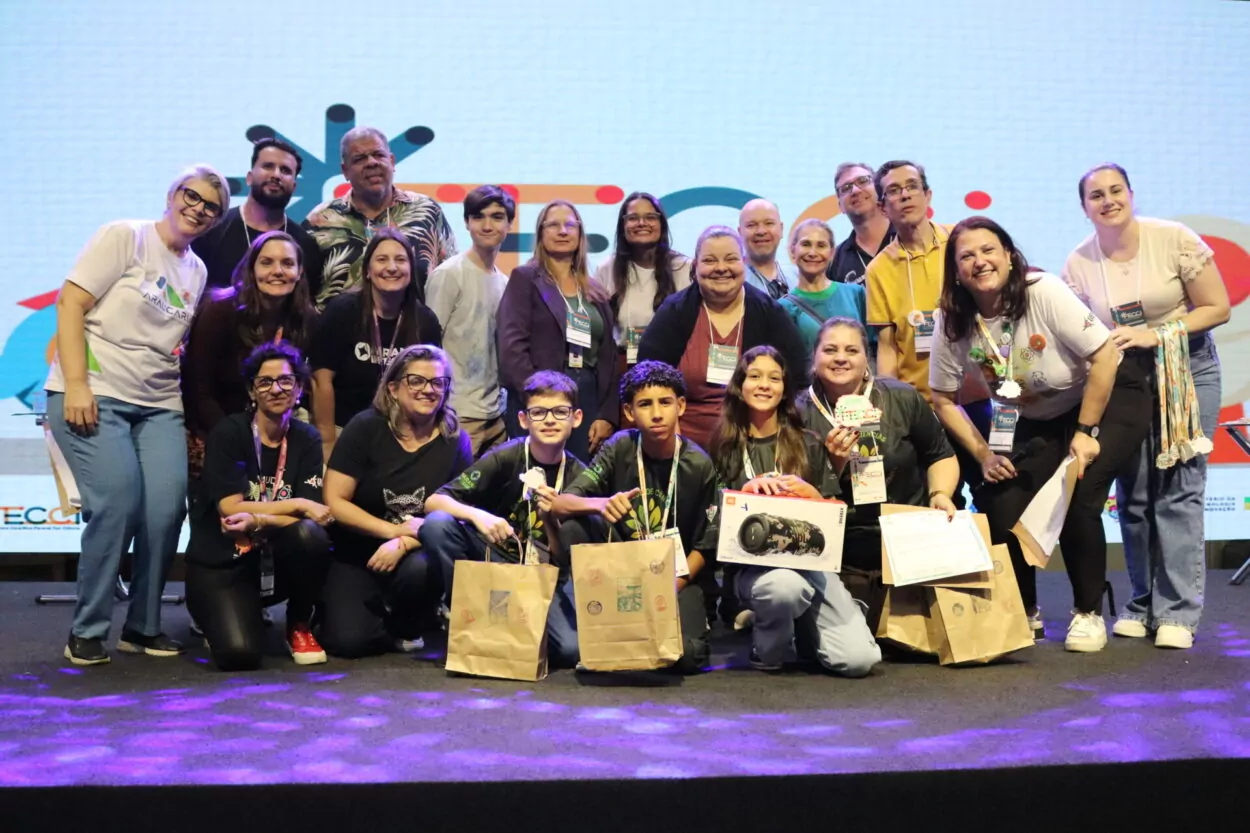 A primeira imagem é do Clube de Ciências Tenda Cultura Viva, do Colégio Estadual Professora Hilda Trautwein Kamal. Na foto há um grupo de pessoas reunidas sobre um palco, sorrindo e posando para a foto durante um evento. Ao fundo, há um telão sugerindo um encontro educacional. Os participantes estão lado a lado, segurando certificados, sacolas de papel e prêmios. Uma pessoa ao centro segura uma grande caixa de uma caixa de som JBL. Todos usam crachás de identificação e vestem roupas casuais. Na segunda fotografia vemos o Dengue Bot, do Colégio Estadual Vereador José Balan. Um grupo grande de pessoas reunidas sobre um palco, sorrindo para uma foto coletiva. Ao fundo, há um telão iluminado com elementos gráficos coloridos, sugerindo que o evento é de caráter educacional. Na parte da frente, alguns jovens seguram sacolas de papel e usam crachás de identificação e camisetas com o logotipo do Paraná. O Paisagismo Sustentável da Escola Estadual Manuel Bandeira, de Alto Piquiri, é a terceira foto. A imagem mostra um grupo numeroso de pessoas reunidas sobre um palco, posando para uma foto durante um evento educacional. Ao fundo, há um telão iluminado com elementos gráficos, reforçando o clima festivo e institucional. Na parte da frente, alguns jovens e adultos exibem certificados, troféus e prêmios. A quarta foto é do Clube Green Lab do Colégio Estadual Almirante Tamandaré, de Cruzeiro do Oeste. Na imagem é possível ver pessoas posando juntas em um palco. Há cerca de quinze pessoas, entre adultos e jovens, algumas agachadas na frente e outras em pé atrás. Os jovens na frente seguram certificados e sacolas de papel. No fundo, há um telão grande com um logotipo colorido.