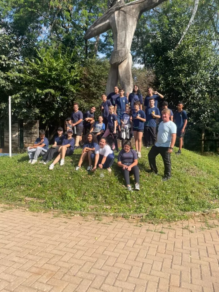 Na primeira fotografia é possível ver um grupo de pessoas posando juntas dentro de uma sala de aula. Ao todo, são dez pessoas: oito jovens e dois adultos. Eles estão posicionados lado a lado. O ambiente é típico de sala de aula, com paredes amarelas, quadro verde ao fundo e piso de cerâmica. A segunda imagem é do Clube Ilha da Ciência. A imagem mostra um grupo de estudantes e duas mulheres posando em um ambiente ao ar livre, com árvores, arbustos e canteiros de horta ao fundo. O grupo está sorridente, alguns em pé e outros agachados na frente. Na terceira fotografia tem um grupo de estudantes reunidos em um ambiente escolar ao ar livre, em frente a um jardim vertical com diversas plantas verdes. Eles estão distribuídos em pé e agachados, posando para a foto. A quarta imagem mostra um grupo de estudantes e um adulto reunidos em um espaço ao ar livre, posando para uma foto em frente a uma grande escultura de uma figura humana com os braços abertos. O grupo está distribuído sobre um pequeno morro coberto de grama, alguns sentados e outros em pé. A quinta imagem é do Clube Experimentando a Ciência: Descobrindo cores e novos sabores. A imagem mostra um grupo de estudantes e professores posando para uma foto ao ar livre, em frente a uma placa de madeira com o desenho de uma abelha e a palavra “Meliponário”. Estão sorridentes e organizados ao redor da placa. Na sexta imagem mostra um grupo grande de pessoas, formado por adolescentes e um adulto, posando para uma foto em uma sala. Eles estão organizados em duas fileiras: alguns sentados ou agachados na frente. A sala tem paredes claras, ventiladores fixados na parede e um quadro branco ao fundo. À esquerda, há uma mesa com uma toalha vermelha e alguns objetos decorativos para um evento escolar. A última fotografia tem um grupo de estudantes e alguns adultos reunidos para uma foto em um ambiente escolar externo. Eles estão em frente a uma parede branca com detalhes verdes e uma janela comprida com grades. O grupo está posicionado em duas fileiras, com alguns agachados na frente e outros em pé atrás, todos sorrindo.