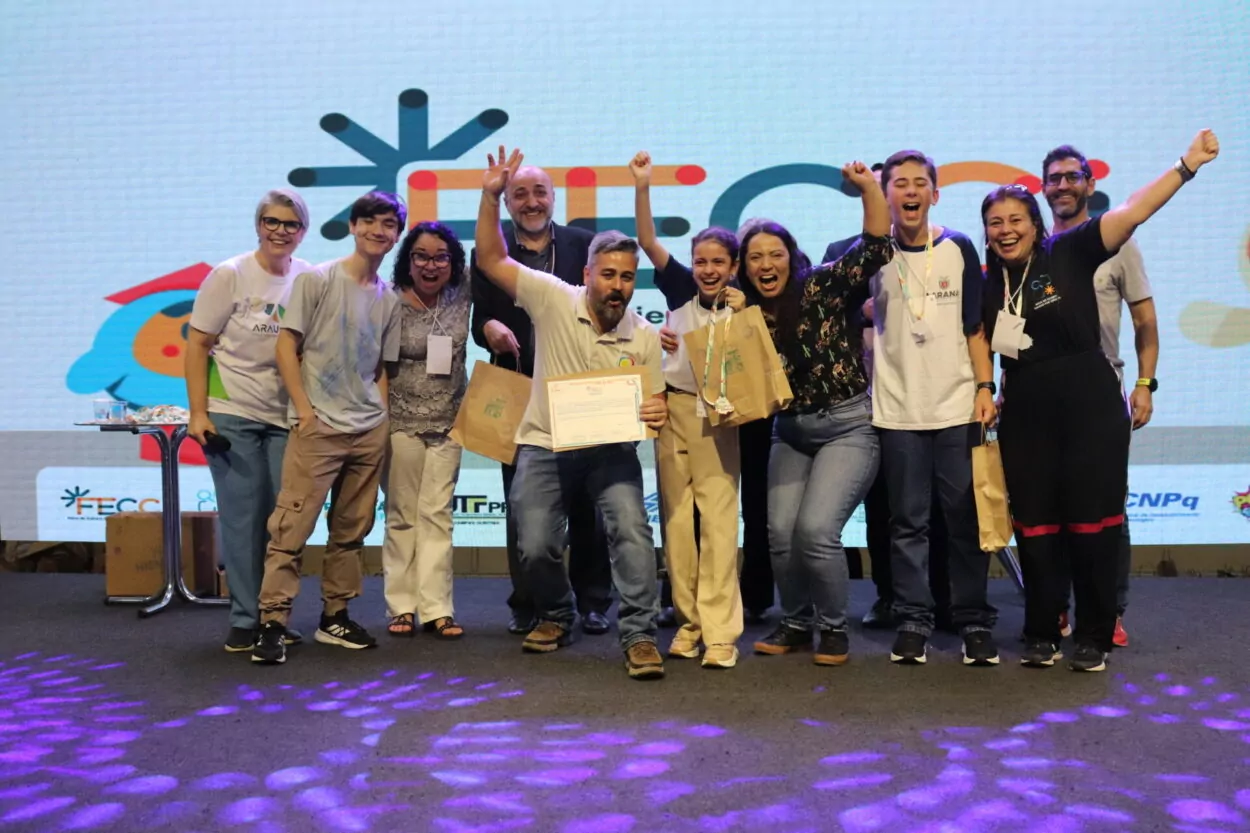 A imagem mostra um grupo de pessoas reunidas em um palco, celebrando com muita alegria. Há cerca de dez pessoas, incluindo adultos e jovens, todas sorrindo, levantando os braços e exibindo entusiasmo. No centro, uma pessoa segura um certificado, que see trata de uma premiaçã. Algumas pessoas também seguram sacolas de papel, que são prêmios do evento O fundo apresenta um grande painel com um logotipo colorido e elementos gráficos, indicando que a foto foi tirada em um evento científico, a FECCI.
