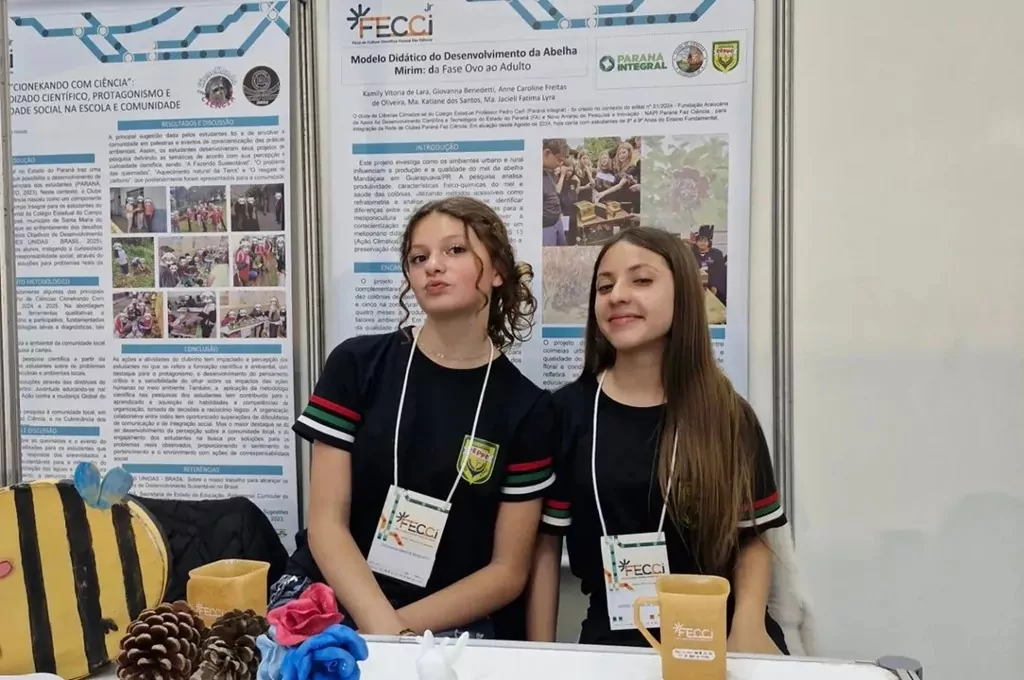 Em uma foto de plano médio, duas jovens estudantes de cabelo castanho claro estão lado a lado em uma feira de ciências. Elas vestem uniformes pretos idênticos e usam credenciais do evento "FECCI". As estudantes estão posicionadas atrás de uma mesa de exposição que contém objetos de projeto, incluindo um modelo de abelha, pinhas e outros itens. Ao fundo, um grande pôster científico branco detalha o projeto delas, com o título "Modelo Didático do Desenvolvimento da Abelha Mirim".