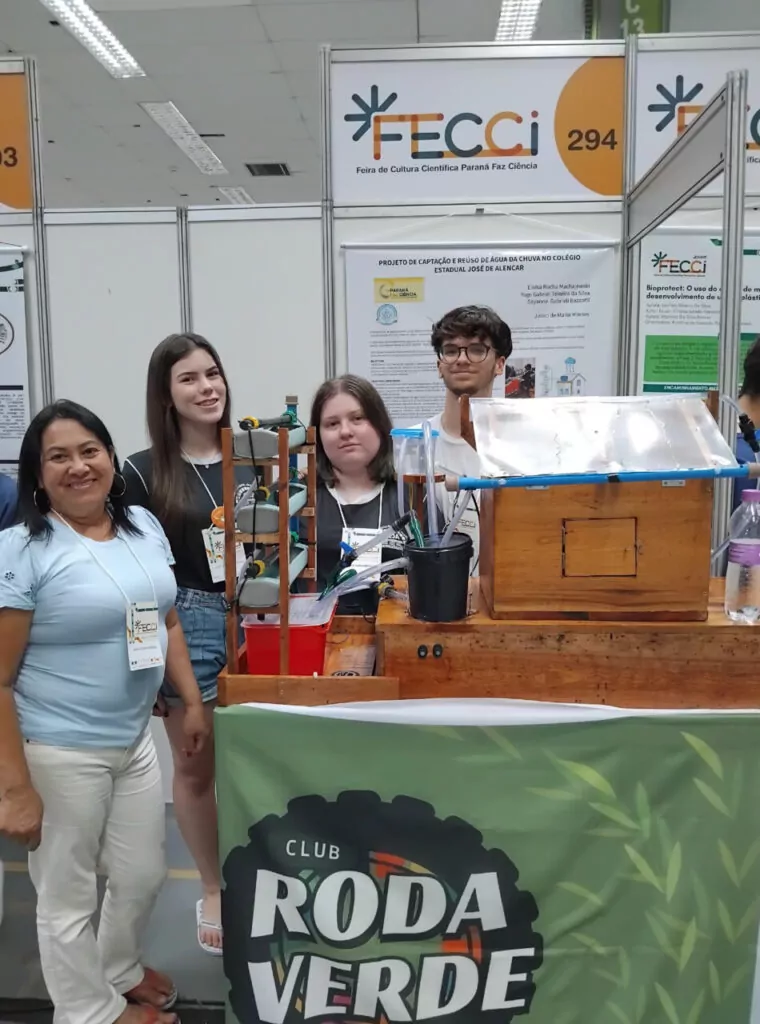 A imagem mostra quatro pessoas em um estande da feira científica FECCI, com um cartaz do projeto “Captação e reuso de água da chuva no Colégio Estadual José de Alencar”, do Clube de Ciências NPI. Elas posam ao lado de uma maquete feita de madeira e tubos que simula o sistema de captação de água da chuva. No fundo, há painéis brancos e o logotipo da FECCI.