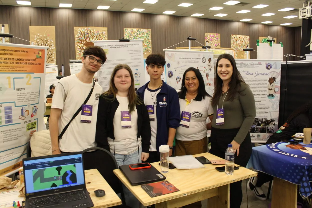 A foto mostra cinco pessoas posando sorridentes em um estande de feira científica. Três estudantes e duas professoras estão lado a lado, com crachás roxos pendurados no pescoço. Atrás deles, há pôsteres de pesquisa com gráficos, textos e ilustrações, identificando projetos de colégios estaduais do Paraná. À frente, sobre uma mesa de madeira, há notebooks, garrafas de água e materiais de apoio. O ambiente é bem iluminado, com várias outras apresentações visíveis ao fundo.