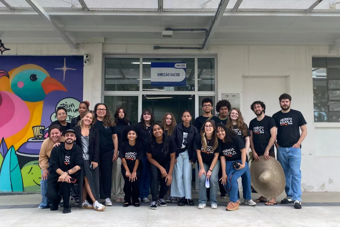 Um grupo de estudantes e integrantes da Agência Escola da UFPR posa sorrindo em frente ao prédio do SACOD, no Campus de Comunicação e Artes da Universidade Federal do Paraná. À esquerda, um mural colorido com desenhos de pássaros compõe o cenário.