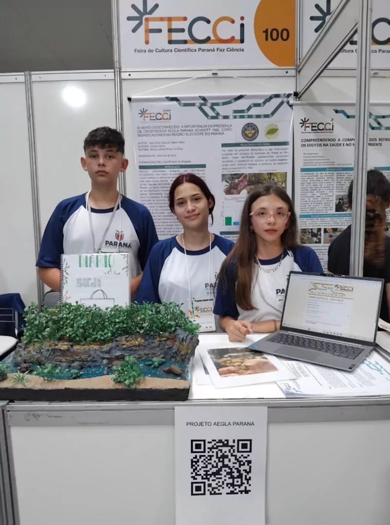 A imagem mostra três estudantes posando atrás de uma mesa na feira científica FECCI, usando camisetas da rede estadual do Paraná. À frente deles, há uma maquete colorida que representa um ambiente natural com vegetação, rio e solo. Um notebook e cartazes com informações complementares estão sobre a mesa. No fundo, um painel apresenta o projeto intitulado “O herói desconhecido: a importância da presença de crustáceos Aegla parana Schimitt 1942, como bioindicadores na região sudoeste do Paraná”, desenvolvido pelo Clube Heróis da Sustentabilidade.