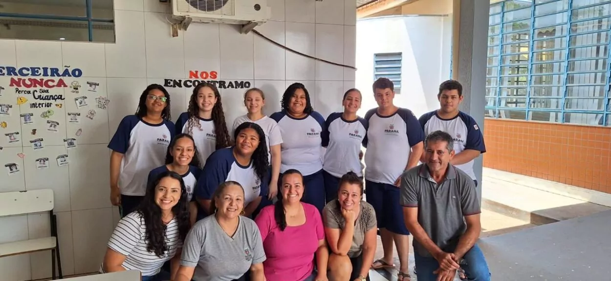 A imagem mostra um grupo de pessoas reunidas em um ambiente escolar. Na frente, há quatro mulheres e um homem agachados, enquanto ao fundo estão mais dez pessoas, entre estudantes e educadores, posando e sorrindo para a foto. Os estudantes vestem uniformes da rede estadual do Paraná, com camisetas brancas e azuis. Ao fundo, há paredes revestidas com azulejos brancos e um mural com frases e desenhos coloridos.