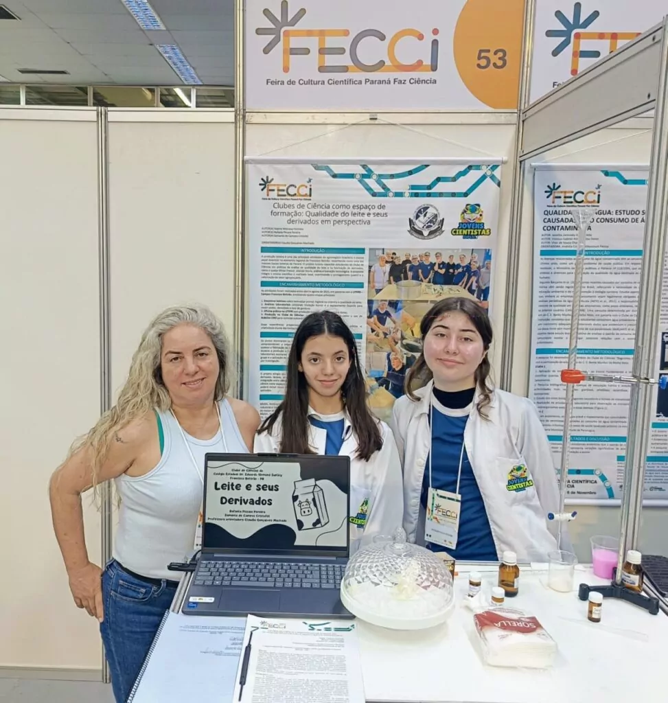 A galeria mostra 5 fotos dos momentos do Clube de Ciências Jovens Cientistas na Feira de Cultura Científica Paraná Faz Ciência (FECCI). O ambiente é coberto e bem iluminado, com estandes e objetos para apresentação da pesquisa desenvolvida pelos clubistas. Há fotos que mostram o banner e os recipientes transparentes nos estandes. A última foto mostra os clubistas em um palco, posando com medalhas, certificados e prêmios pelo 2º lugar na categoria Ciências da Saúde Junior com o projeto “Clubes de Ciência como Espaço de Formação: Qualidade do Leite e seus Derivados em Perspectiva”.