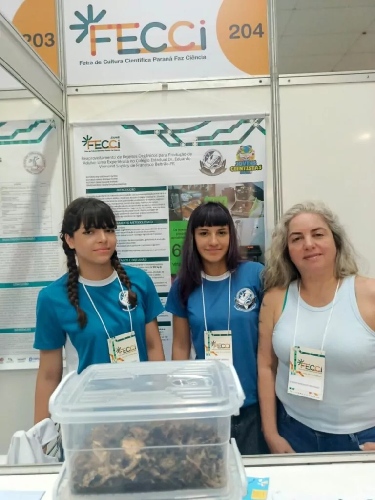 A galeria mostra 5 fotos dos momentos do Clube de Ciências Jovens Cientistas na Feira de Cultura Científica Paraná Faz Ciência (FECCI). O ambiente é coberto e bem iluminado, com estandes e objetos para apresentação da pesquisa desenvolvida pelos clubistas. Há fotos que mostram o banner e os recipientes transparentes nos estandes. A última foto mostra os clubistas em um palco, posando com medalhas, certificados e prêmios pelo 2º lugar na categoria Ciências da Saúde Junior com o projeto “Clubes de Ciência como Espaço de Formação: Qualidade do Leite e seus Derivados em Perspectiva”.