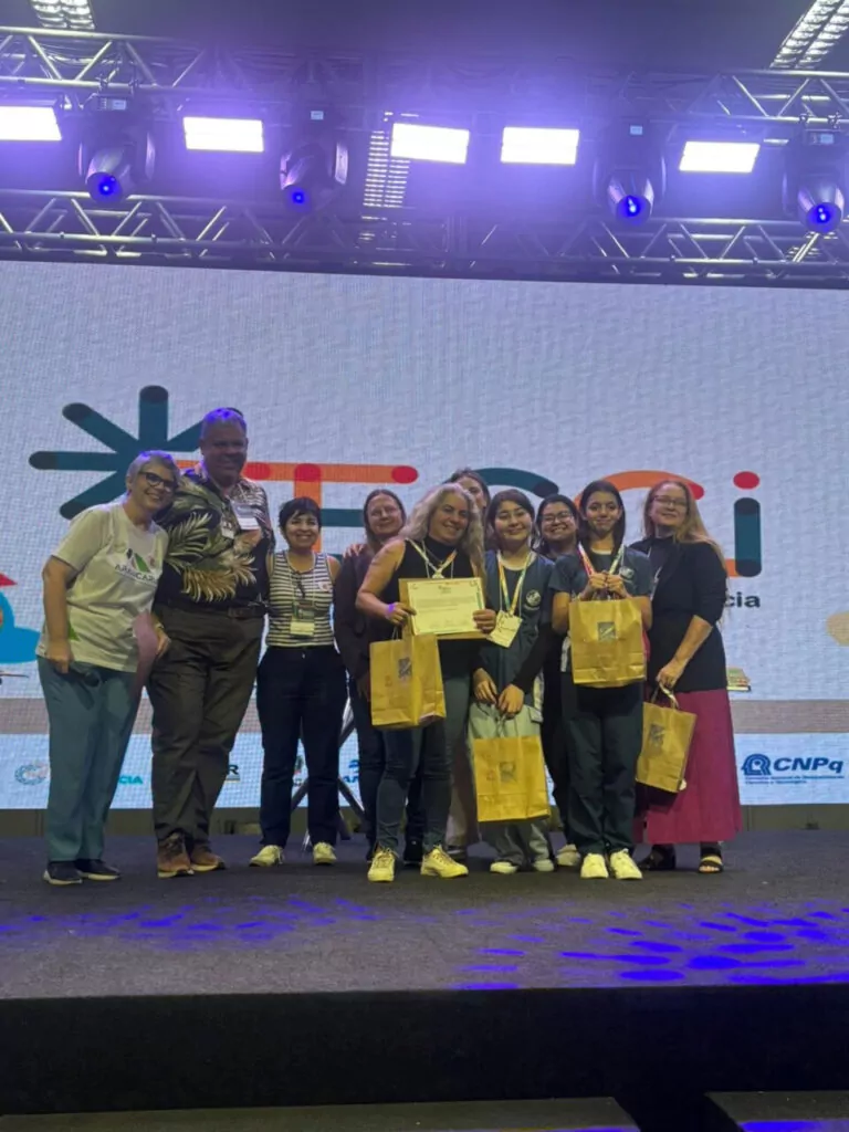 A galeria mostra 5 fotos dos momentos do Clube de Ciências Jovens Cientistas na Feira de Cultura Científica Paraná Faz Ciência (FECCI). O ambiente é coberto e bem iluminado, com estandes e objetos para apresentação da pesquisa desenvolvida pelos clubistas. Há fotos que mostram o banner e os recipientes transparentes nos estandes. A última foto mostra os clubistas em um palco, posando com medalhas, certificados e prêmios pelo 2º lugar na categoria Ciências da Saúde Junior com o projeto “Clubes de Ciência como Espaço de Formação: Qualidade do Leite e seus Derivados em Perspectiva”.